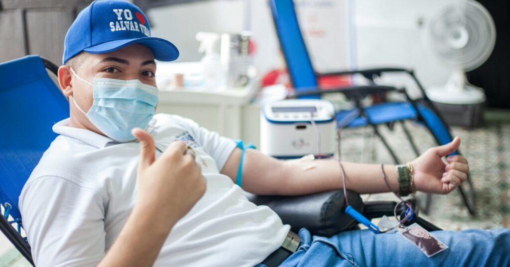 リクライニングチェアに座り、献血をするマスクと青い帽子をかぶった男性。傍らには医療器具が見える。.